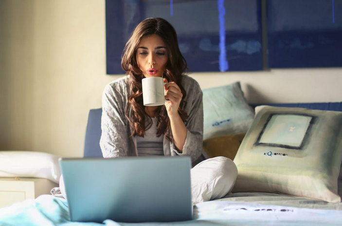 Woman sitting on a bed, looking at a laptop and holding a cup of coffee in her hand